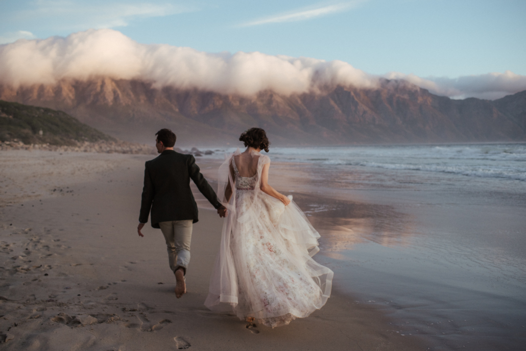 A couple walking along a Cape Town beach with mountains and clouds at sunset